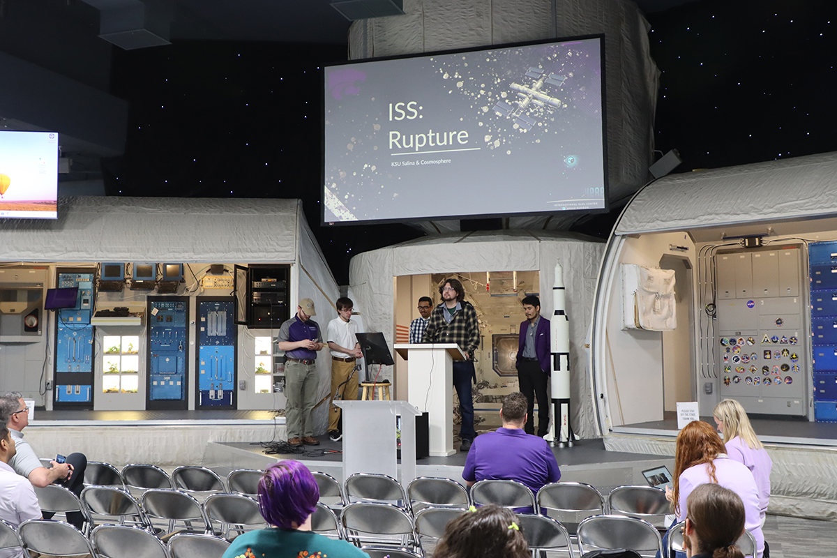 A group of college students stand in a museum's large hall and present about a virtual reality space simulation game they developed.