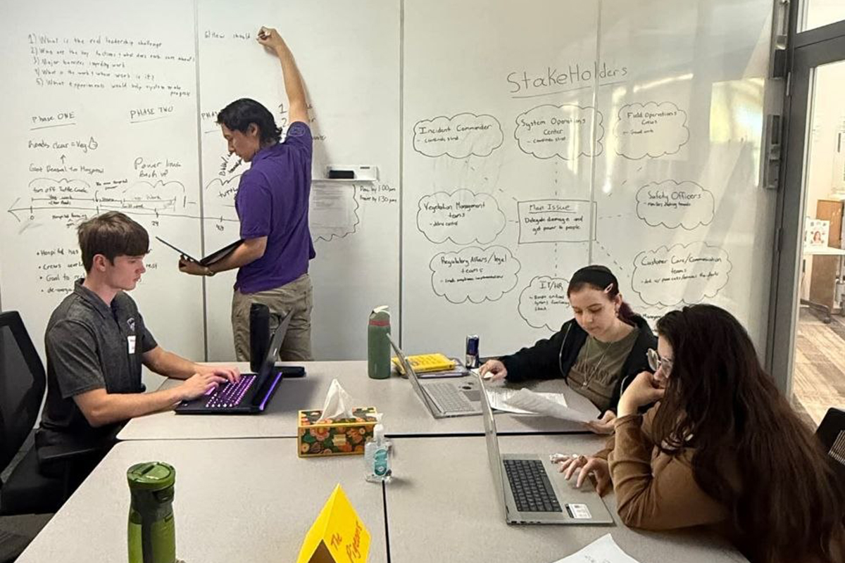 A group of people sit in a small study room and write on a whiteboard on a wall.