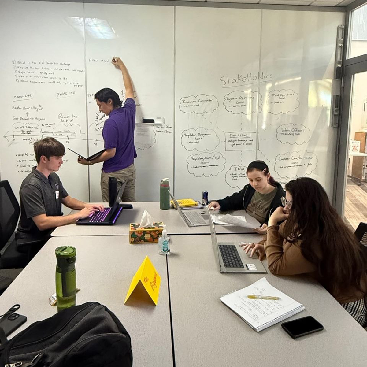 A group of people sit in a small study room and write on a whiteboard on a wall.