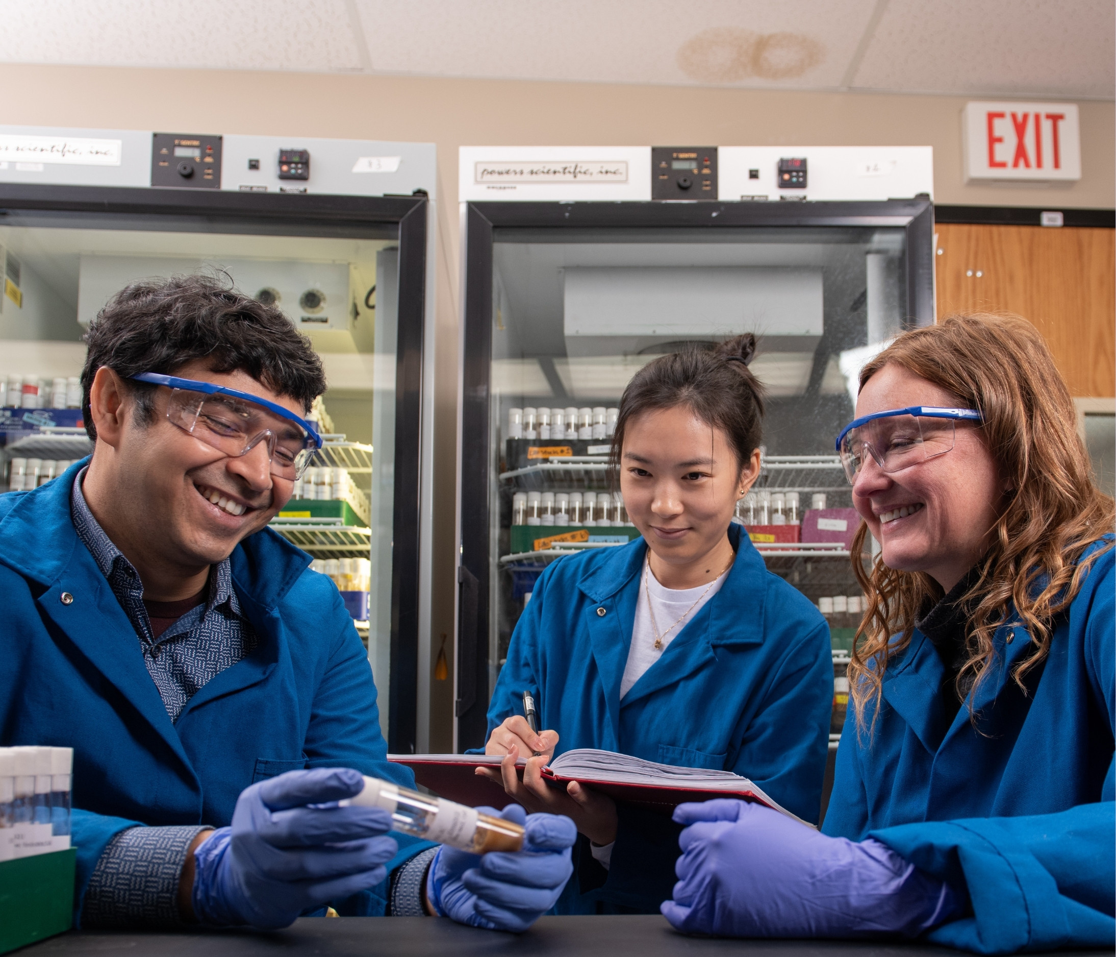 Geisbrecht analyzes lab samples with two students. All three wear blue lab coats and goggles. 