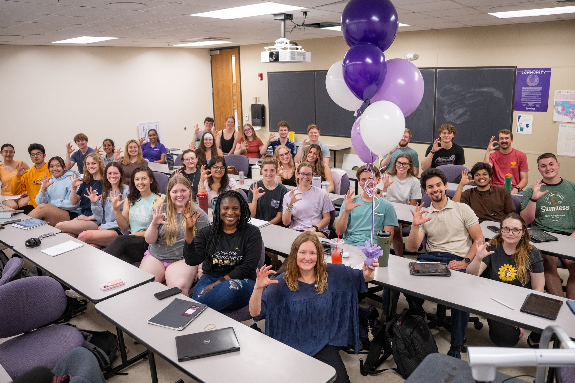 Geisbrecht poses in a classroom filled with students, holding purple balloons and making a Power Cat hand sign.