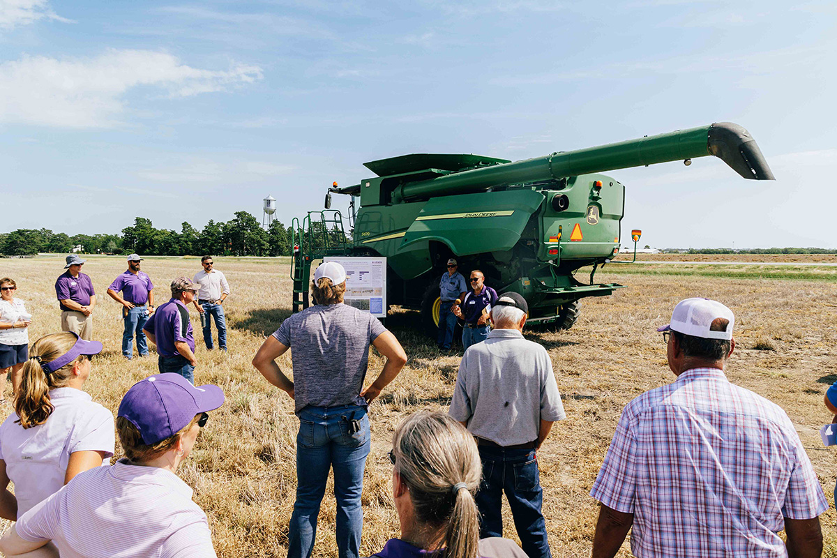 About 15 people stand in the middle of a field near a green combine