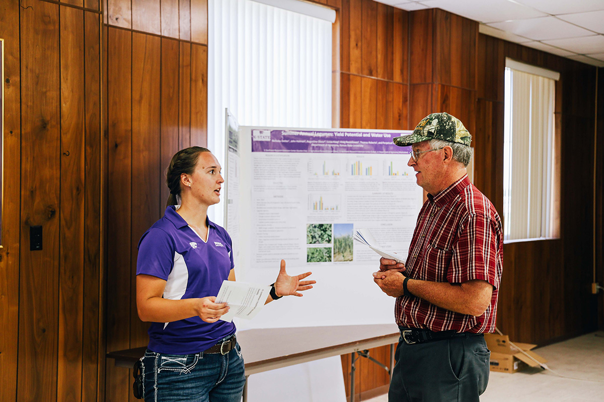 A woman in a purple polo stands next to a research poster and talks to a man wearing a camo hat.