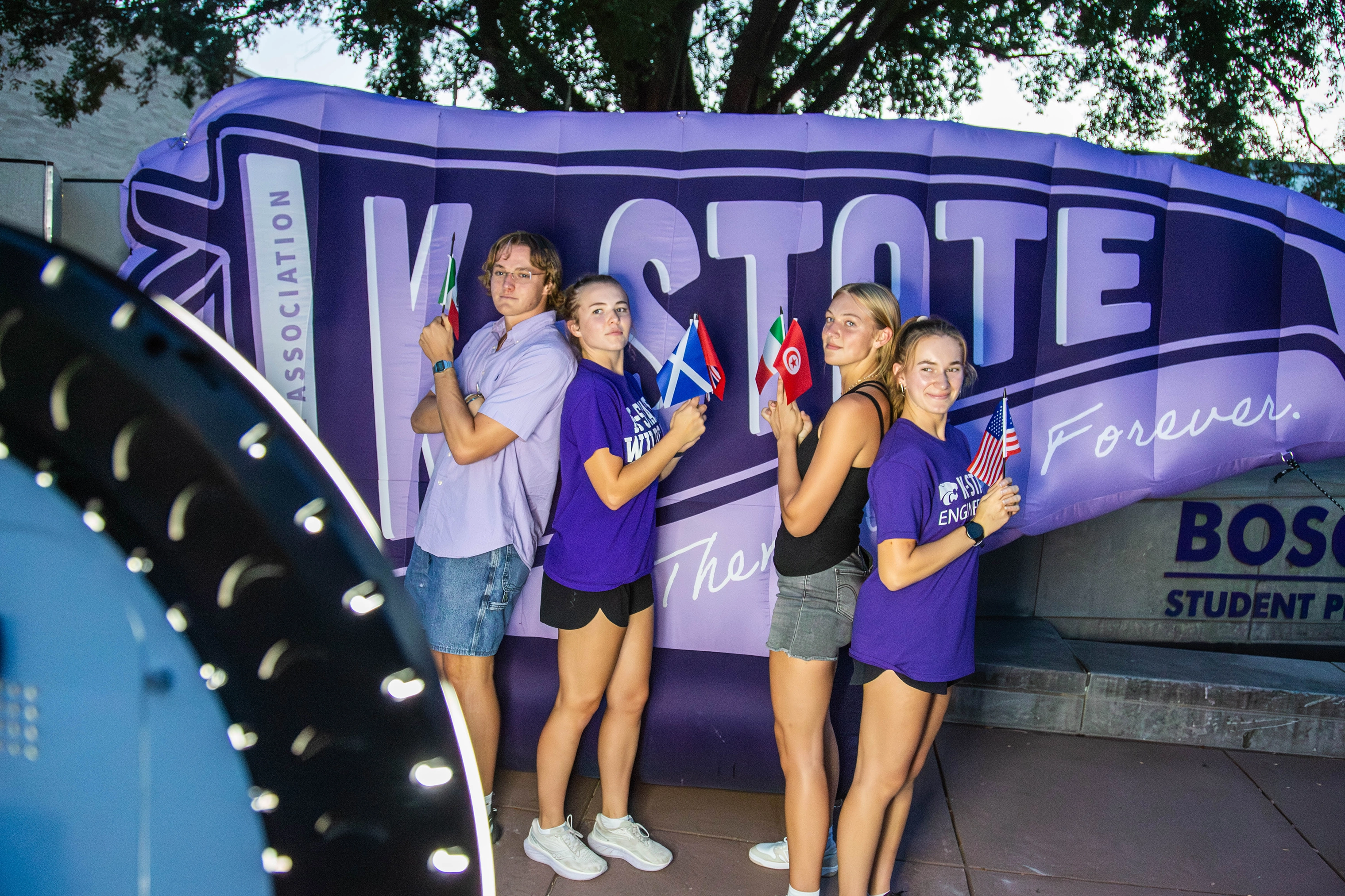 Four students posing in front of an inflatable flag with “K-State” written across the flag. 
