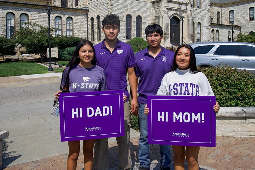 Two girls in front of two guys that are standing in front of Anderson Hall. The girl on the left is holding a sign that says “hi dad” and the girl on the right holding a sign that says “hi mom."