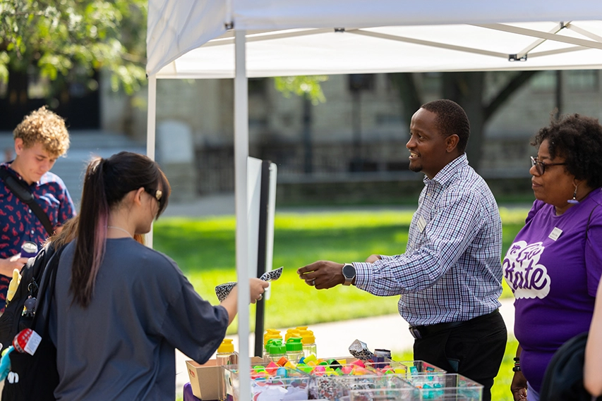 A K-State staff member handing a goodie to a student outside, under a tent.