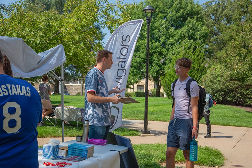 Two individuals talking at a table outside, near a tent.