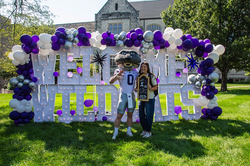 Willie the Wildcat posing with a student in front of giant letters that spell out “Welcome Wildcat”. The female student is holding a sign that says, “Happy 1st Day of Classes.”