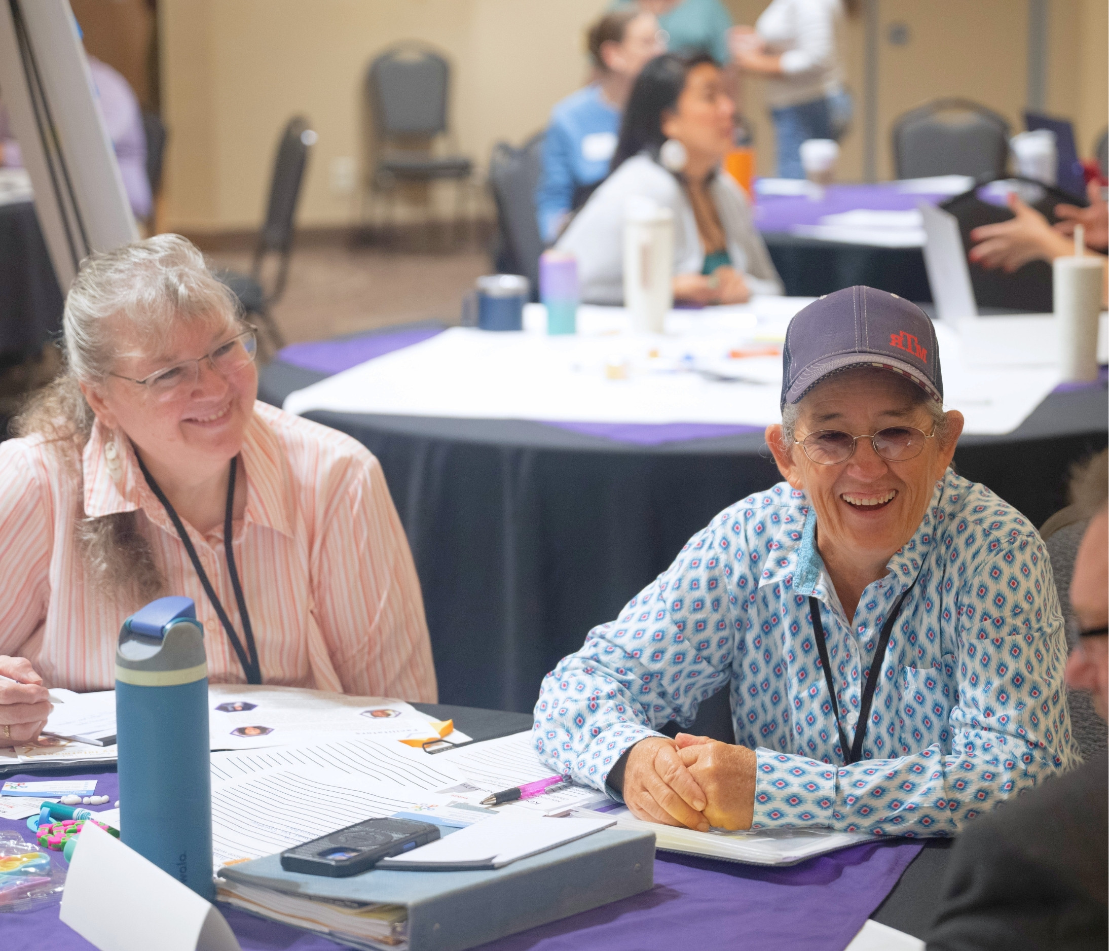Two women smile and participate in a discussion during a business summit, seated at a table with papers and notebooks. Other attendees work together at tables in the background.