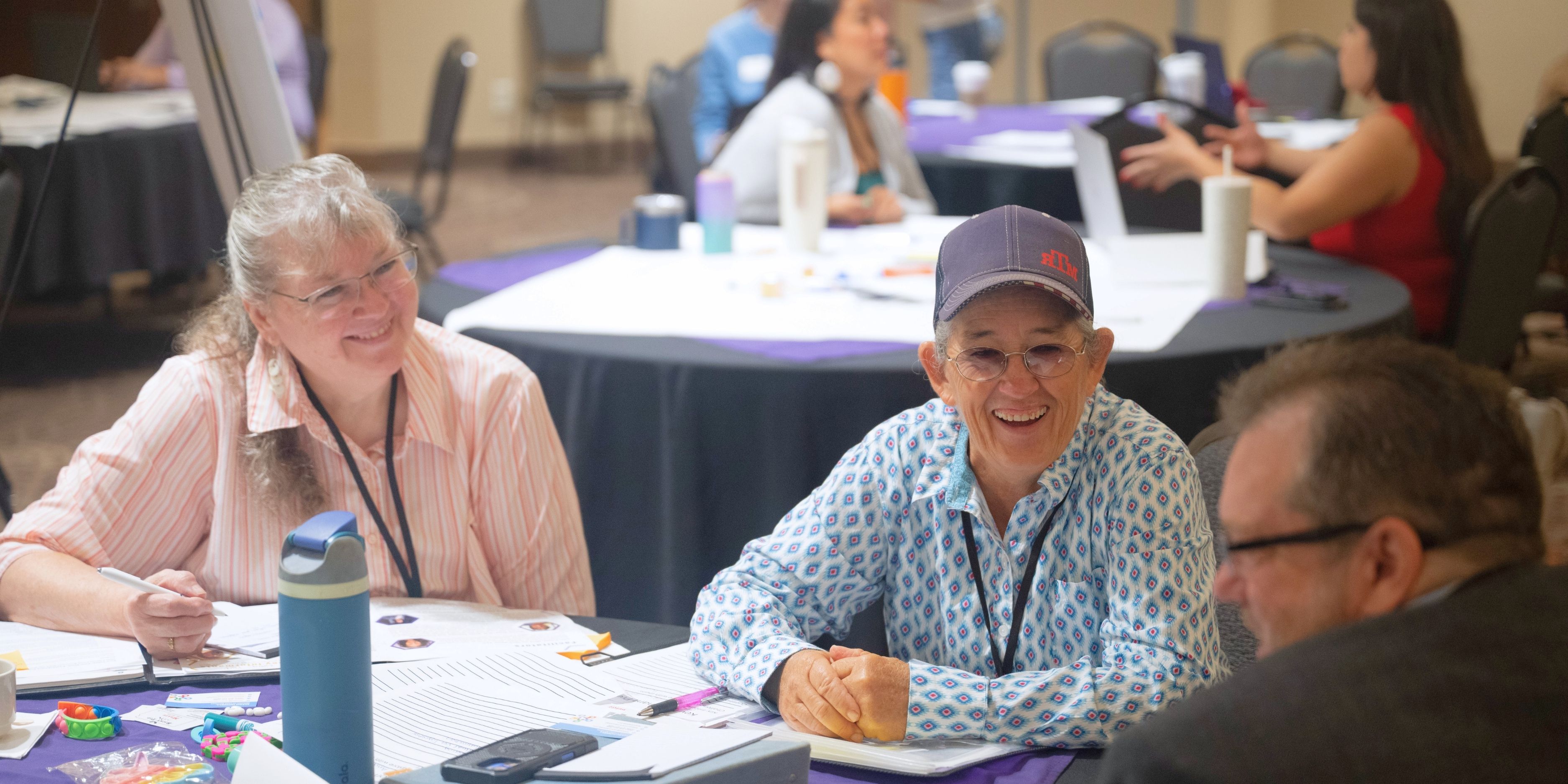 Two women smile and participate in a discussion during a business summit, seated at a table with papers and notebooks. Other attendees work together at tables in the background.