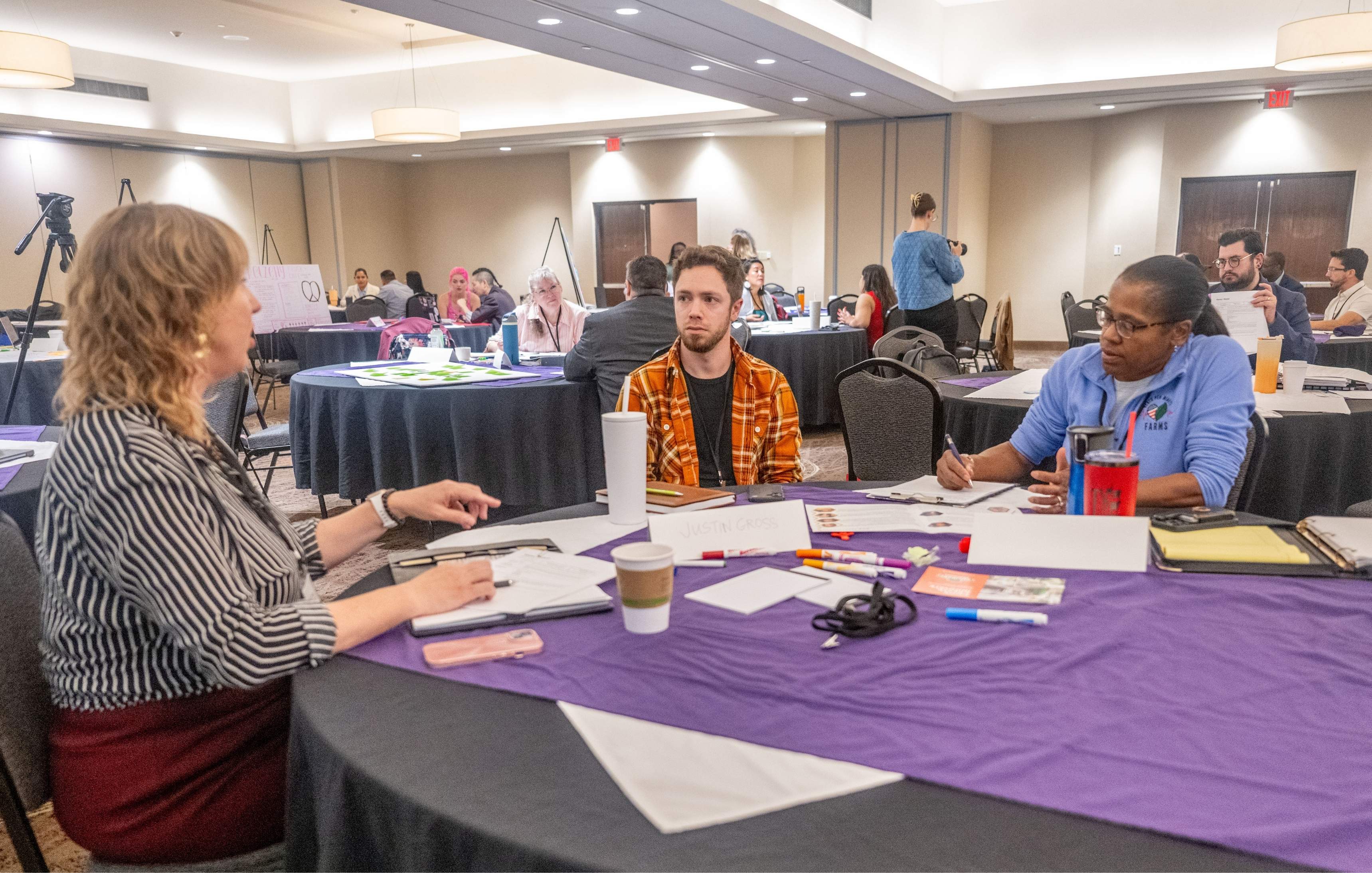 Three business professionals sit at a round table in discussion. On the tablea are a variety of markers and printed workshop materials