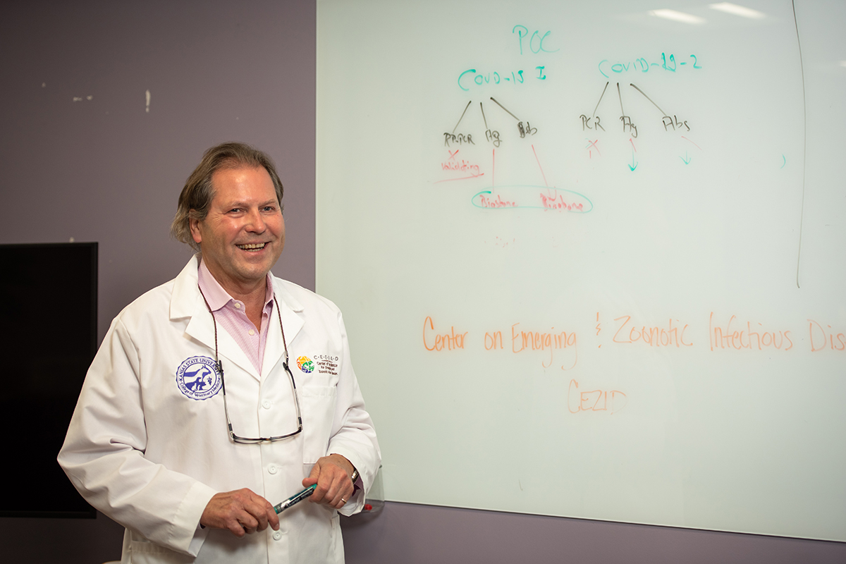 A university researcher in a lab coat poses for a portrait in front of a white board that has written on it 