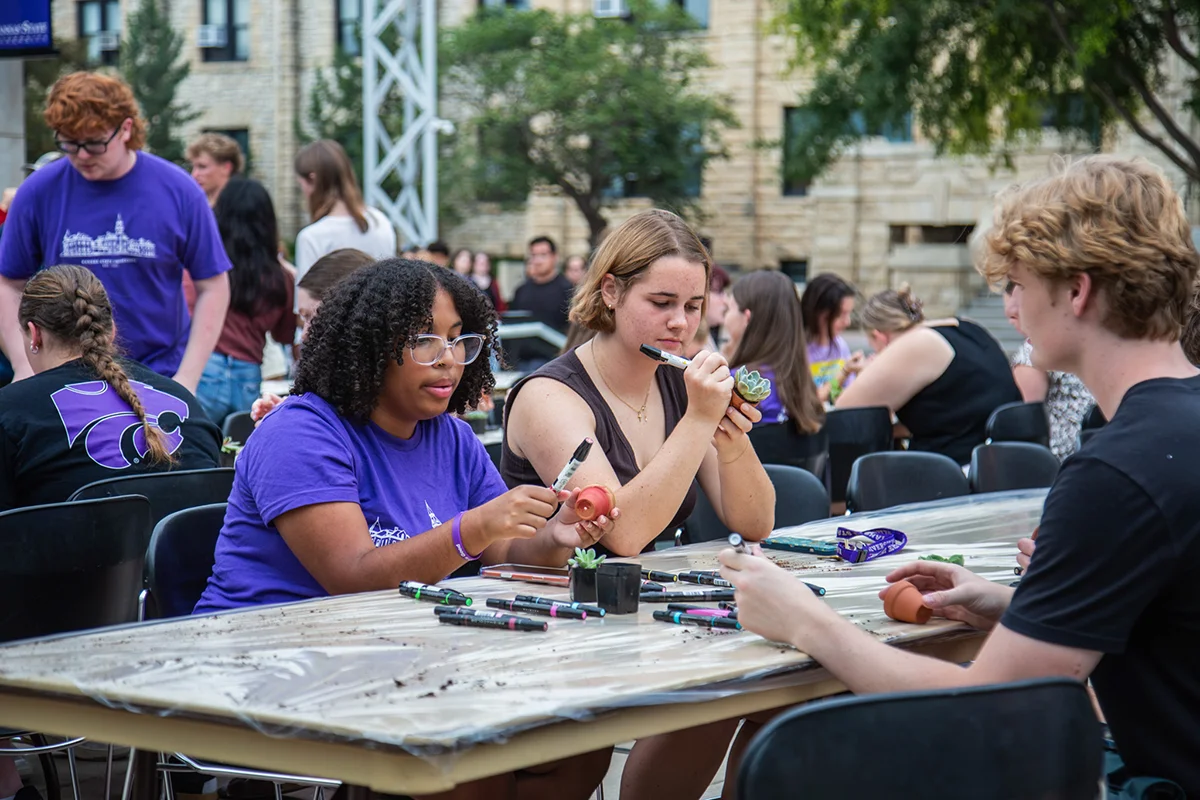 Three students sitting at a long table outside while decorating flowerpots. 