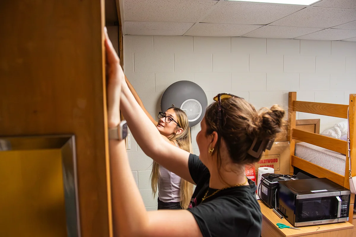 Two students holding up an item in the closet of their dorm room.