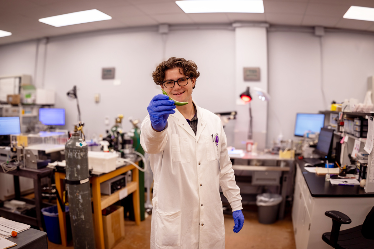 A graduate student in a white lab coat holds a green pepper up for the camera.
