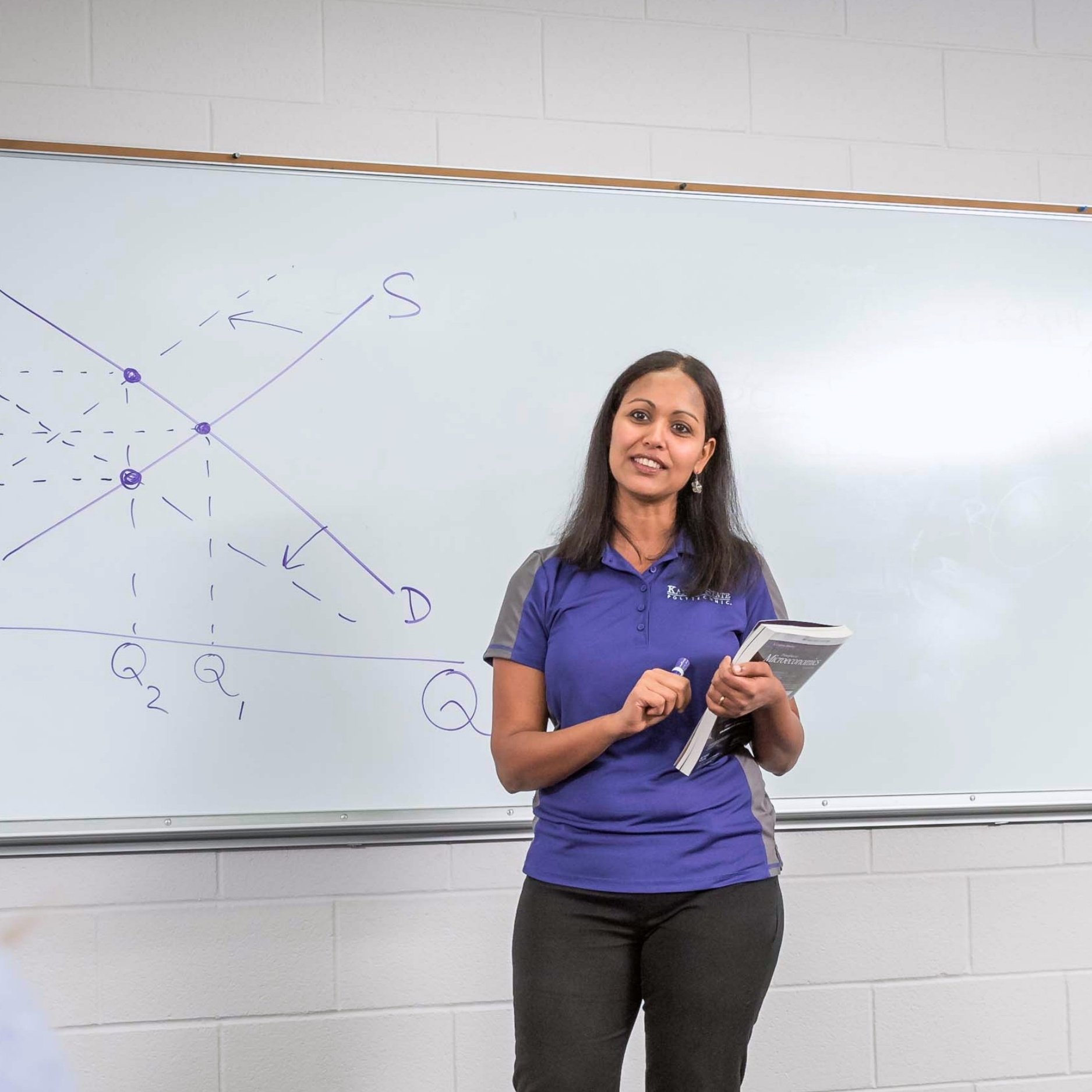Siny Joseph, wearing a purple and grey K-State polo shirt, stands smiling in front of a white board. The board showcases a supply-and-demand model graph written in purple marker.