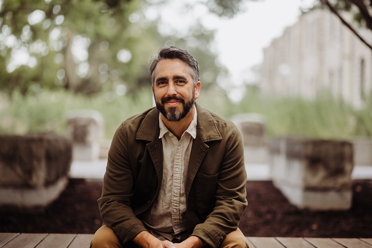 A man poses for an outdoor portrait in front of a limestone college academic building.