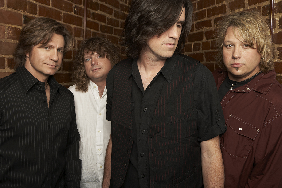 Four men with long hair stand against a brick wall for a band photo.