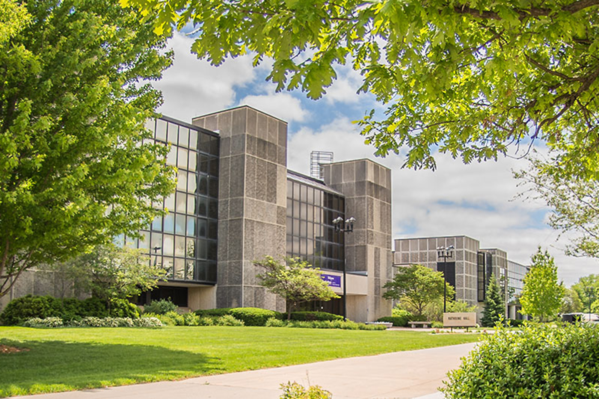 A gray concrete college academic building peaks out from beneath green trees.