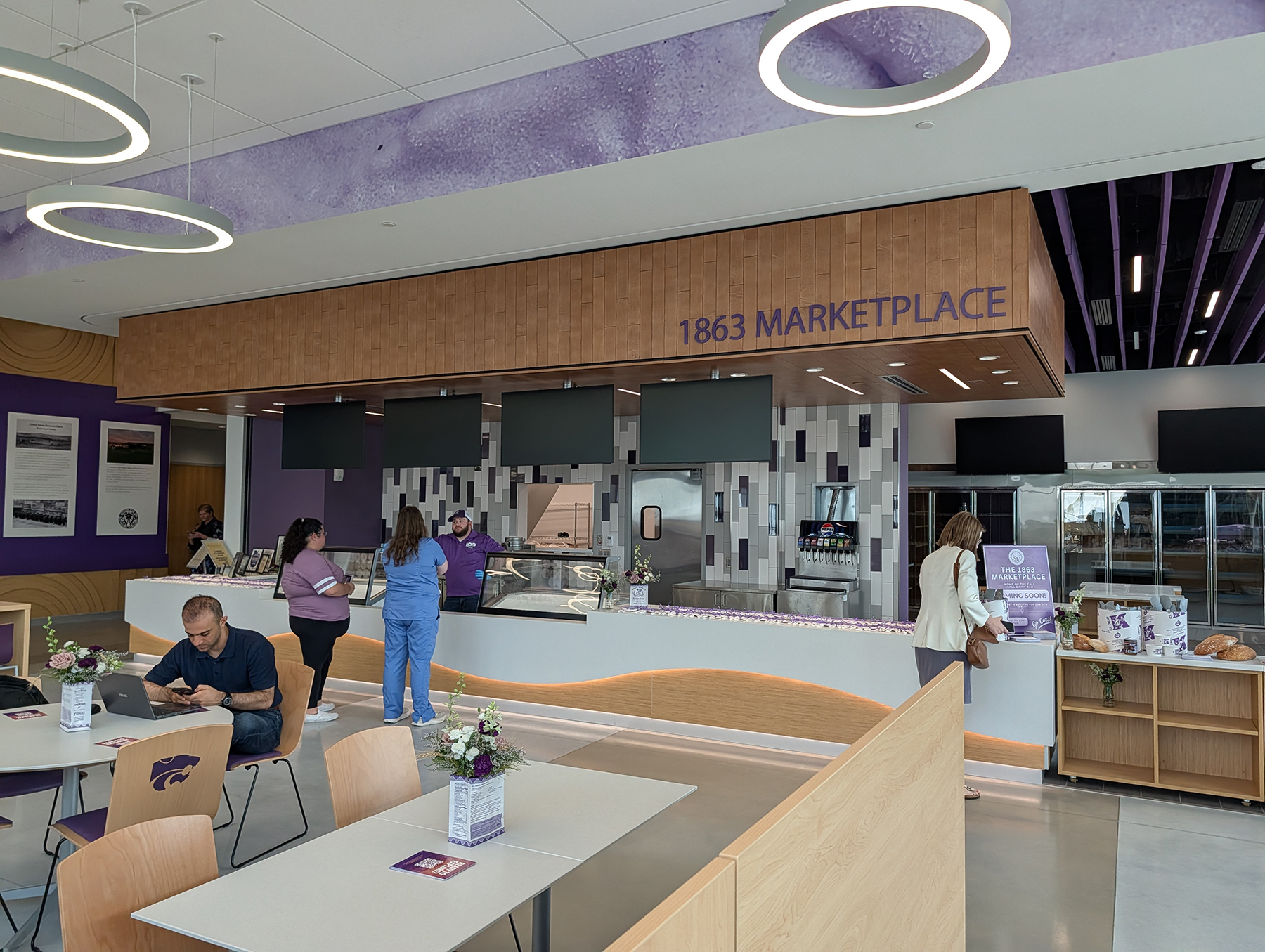 A rendering shows several people waiting in line at a lunch counter inside a modern academic building.