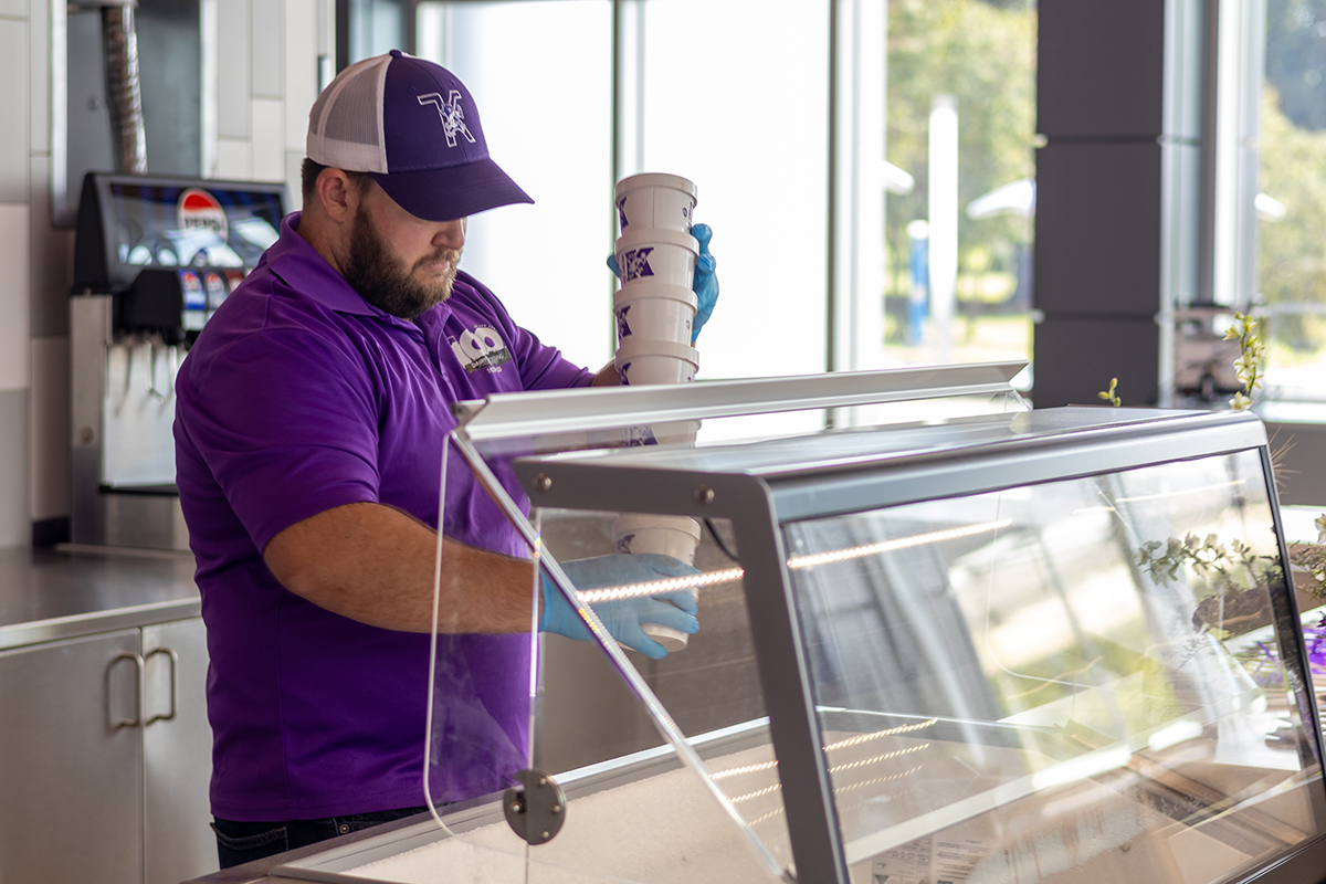 A man carries a stack of paper cups containing ice cream to a cold chest at a store front.