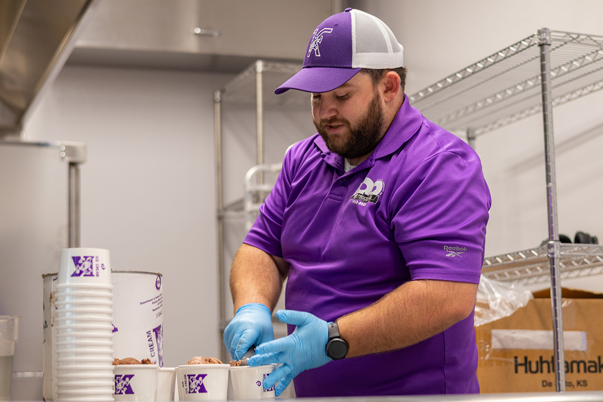 A man in a kitchen serves scoops of chocolate ice cream into paper cups branded with a purple K.