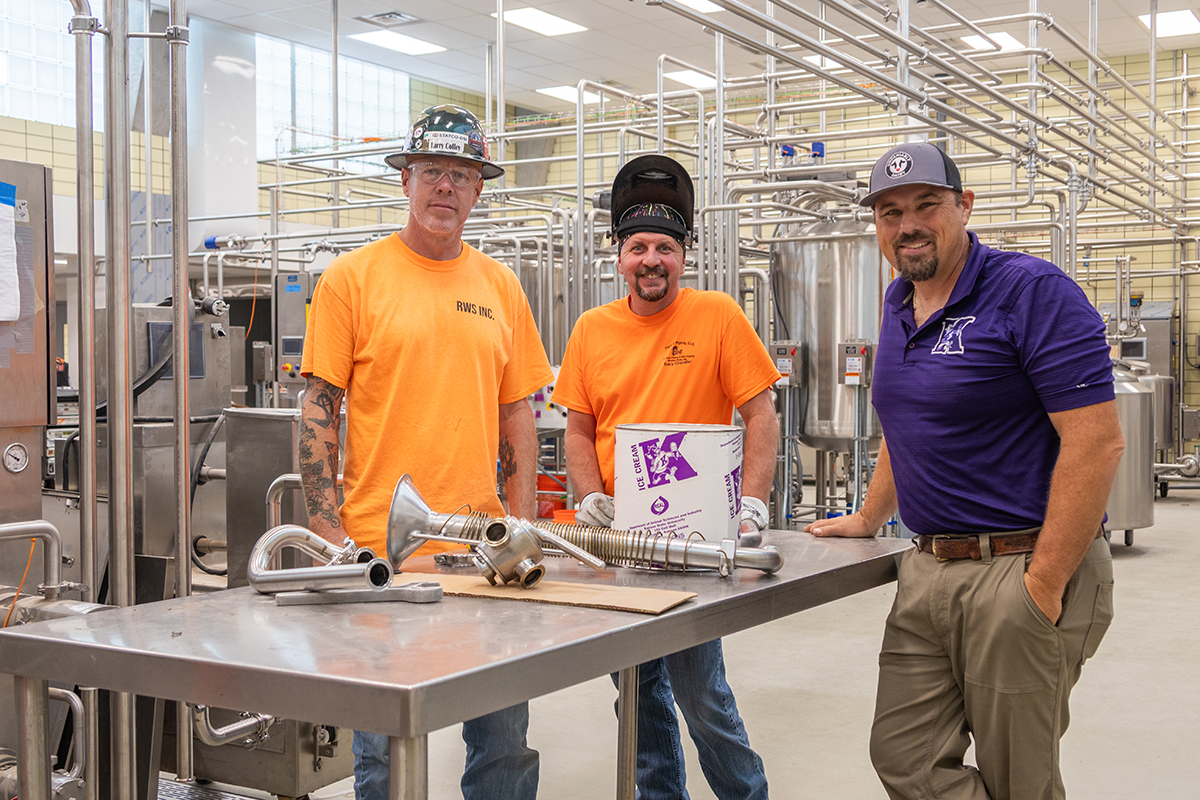 Three men pose for a portrait at a metal table in a dairy plant.