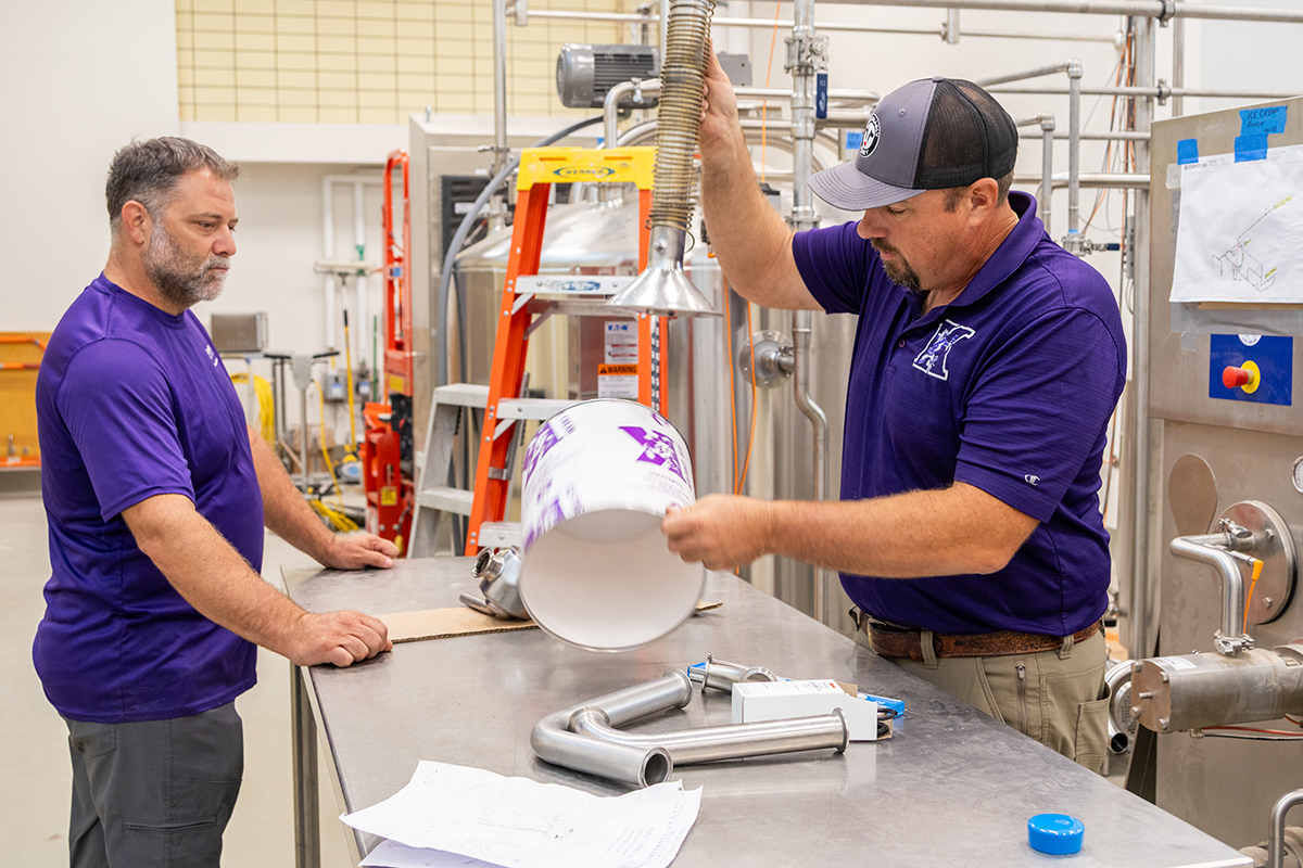 A man demonstrates how a metal tube connects to a large, sturdy paper ice cream bucket in a dairy plant.