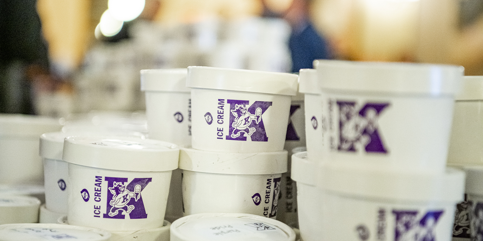 Several white ice cream tubs with a purple letter K on each sit on a table.