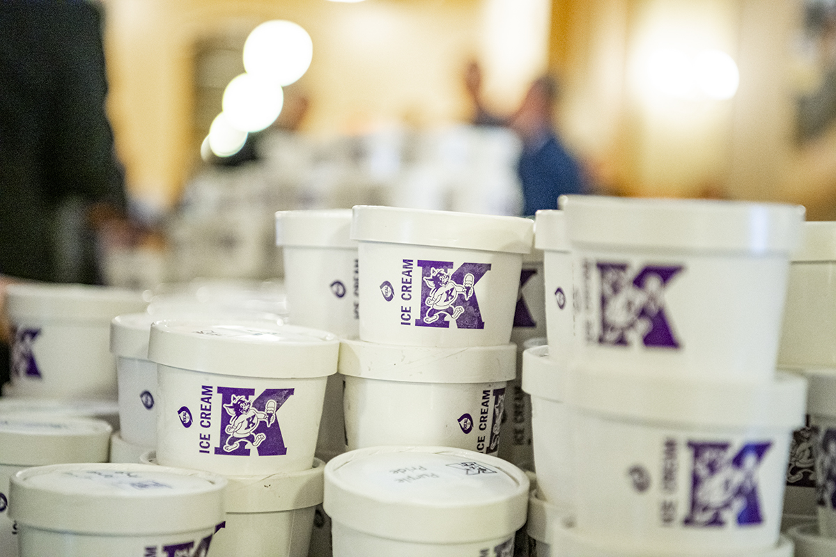 Several white ice cream tubs with a purple letter K on each sit on a table.
