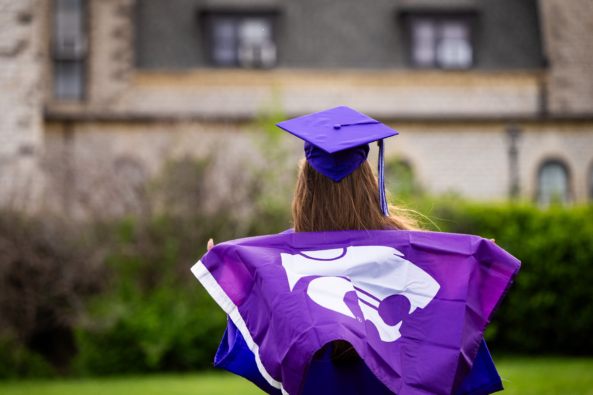 A college student stands facing Anderson Hall at K-State and wears a purple cap and gown and has a purple Powercat flag draped over her shoulders. 