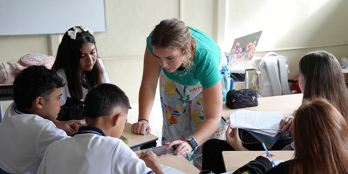 A female college student leans over students' desks and assists them with a classroom activity.