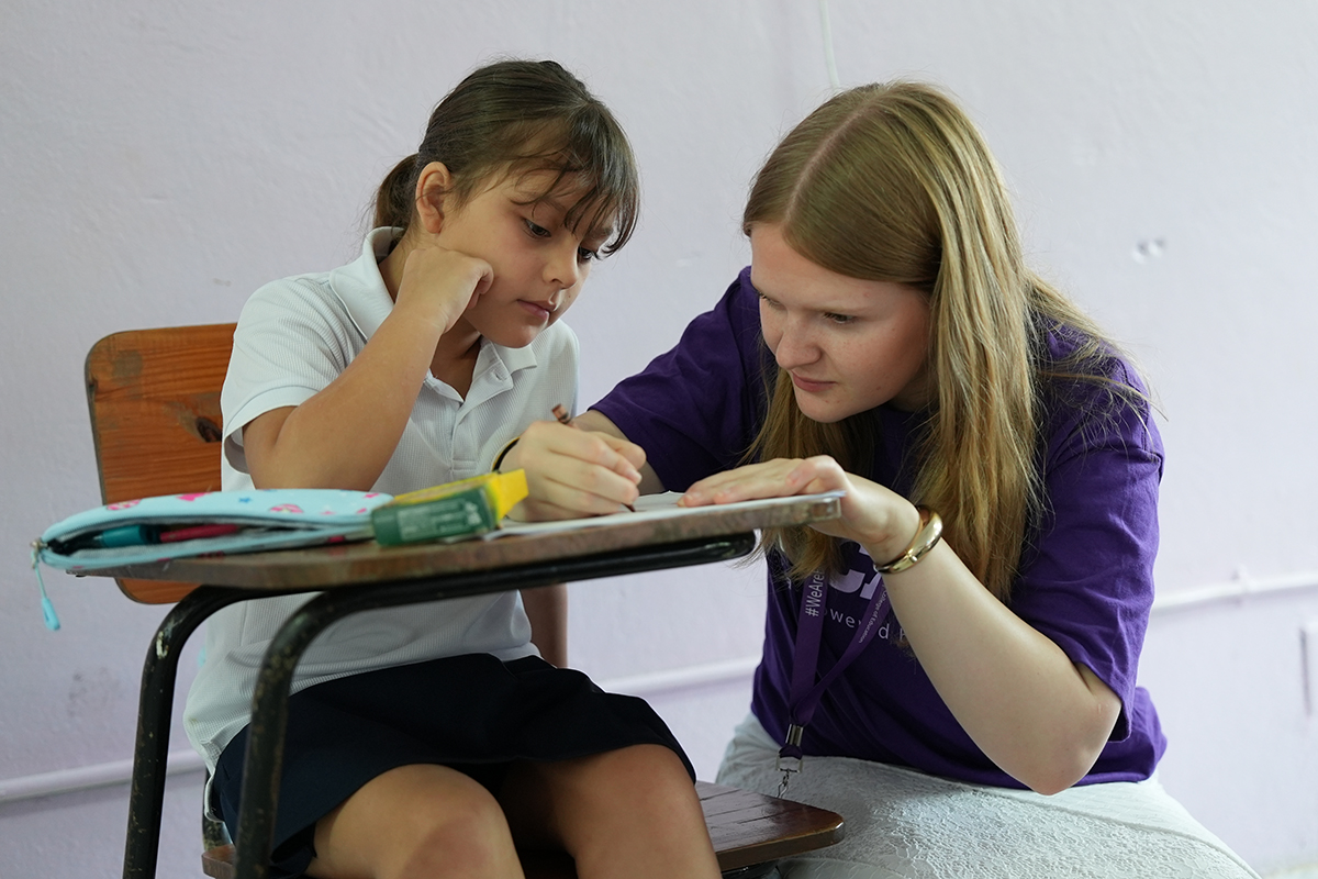 A college education student leans over a Costa Rican students' desk and assists her.