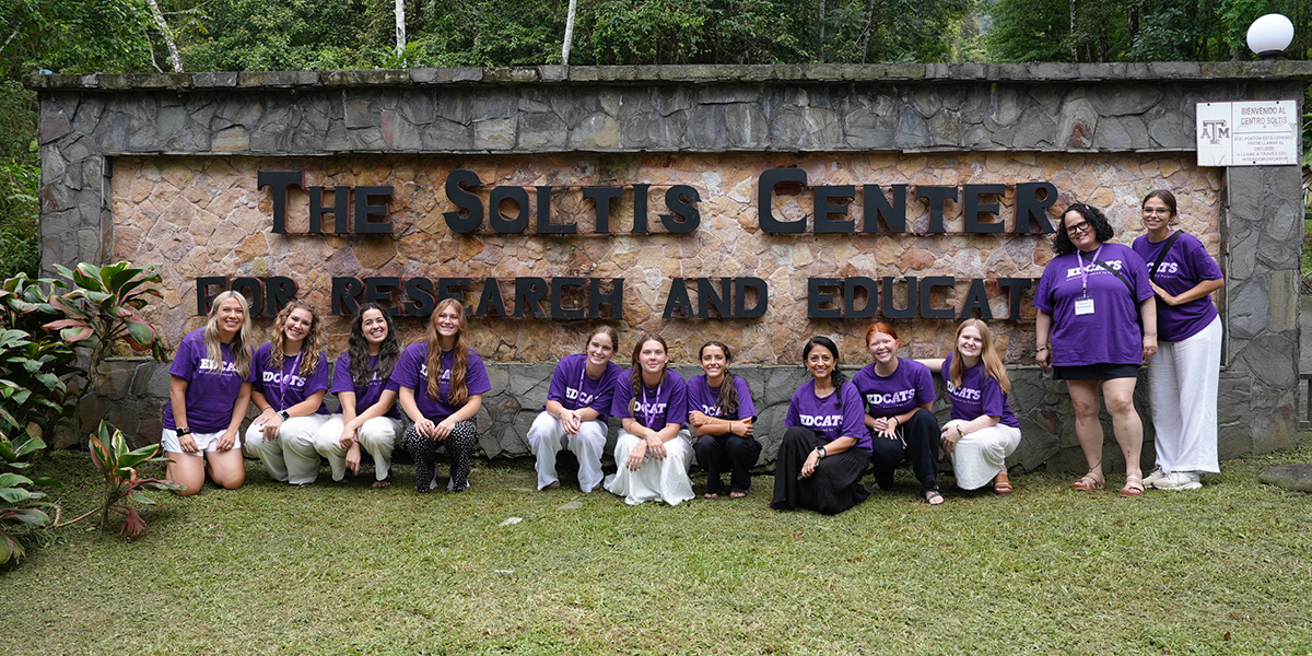 A group of college students pose for a portrait outside a stone sign in a jungle.