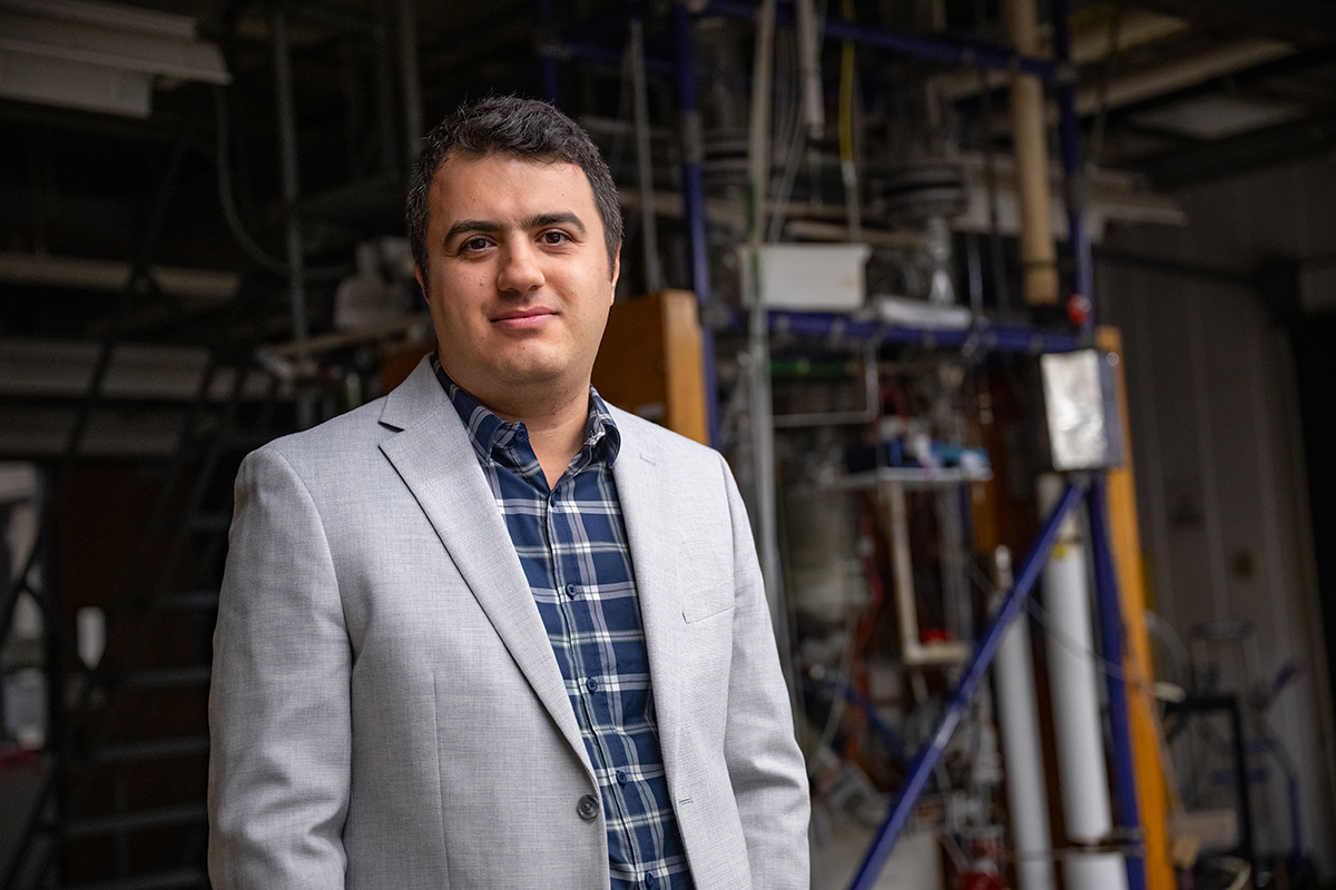A man in a gray suit poses for a portrait in front of various tubing and wires and equipment behind him.