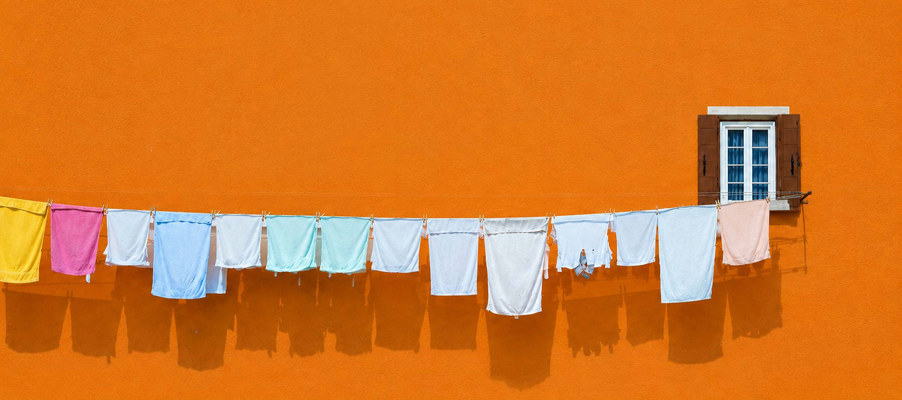 Several pieces of laundry hang from a clothesline against a bright orange wall and a brown-framed window.