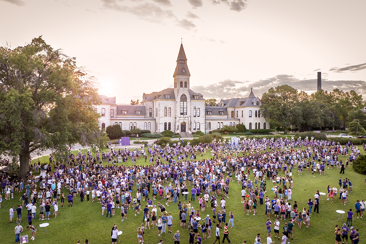 A large crowd of students in mostly purple attire gathers on a lawn in front of a large, towered limestone academic building. The sun is setting behind it.