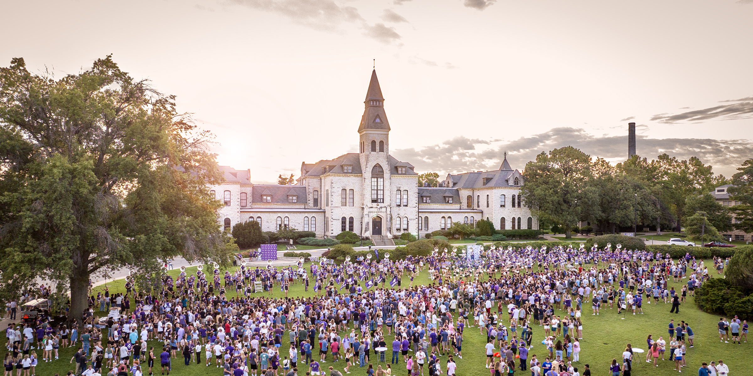 A large crowd of students in mostly purple attire gathers on a lawn in front of a large, towered limestone academic building. The sun is setting behind it.