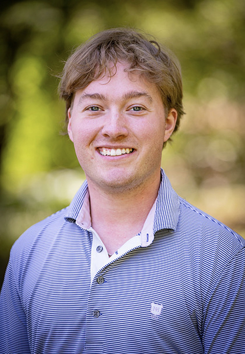 A young man in a striped polo is pictured with a blurred outdoor background.