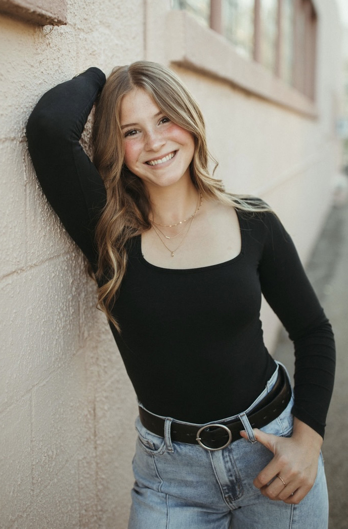 A young woman in a black shirt leans against a wall for a photo.