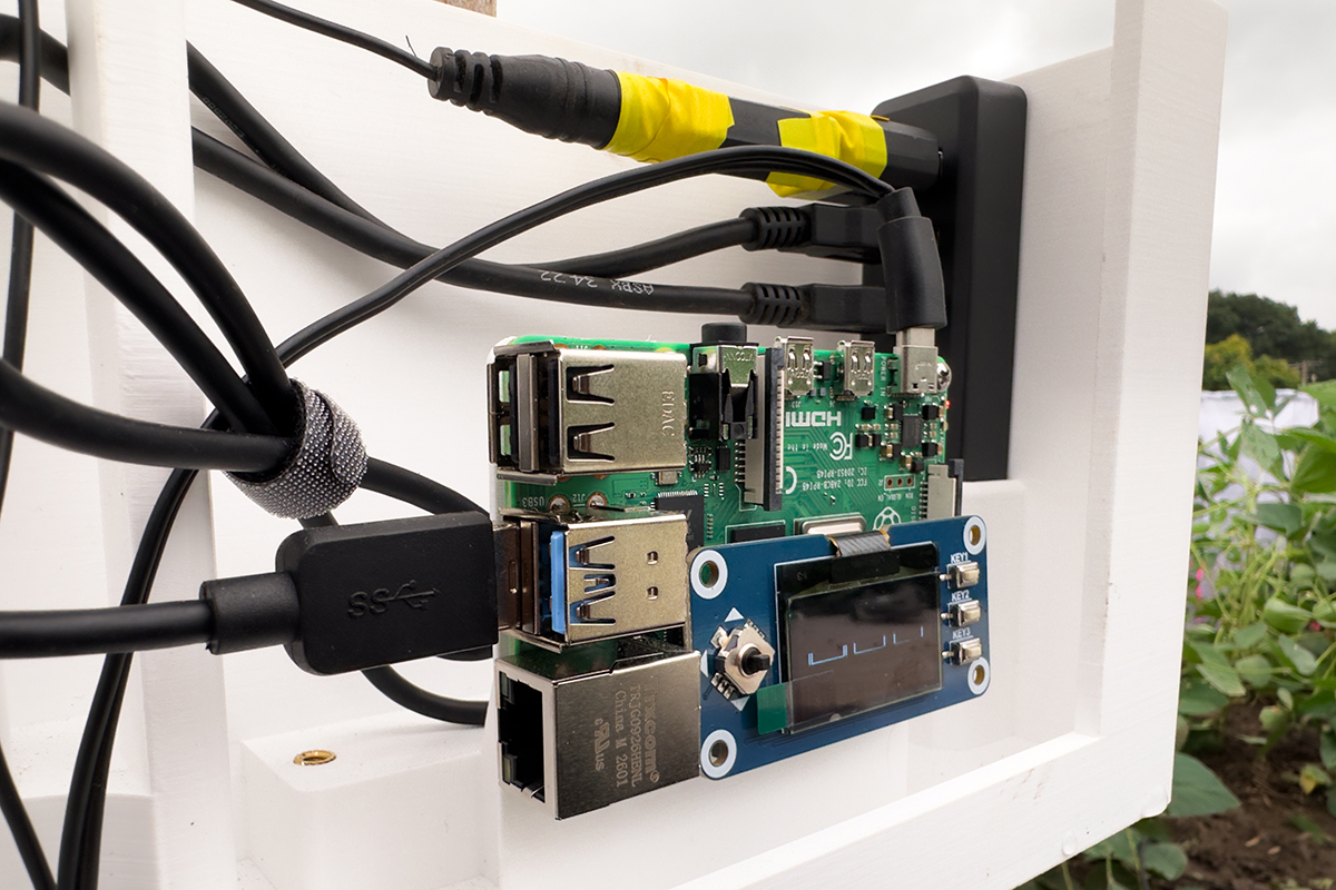 White 3D printed box holds green circuit board with mini screen interface. There are multiple different plug ports with wires leading out of the white casing. Soybeans are in the distance.