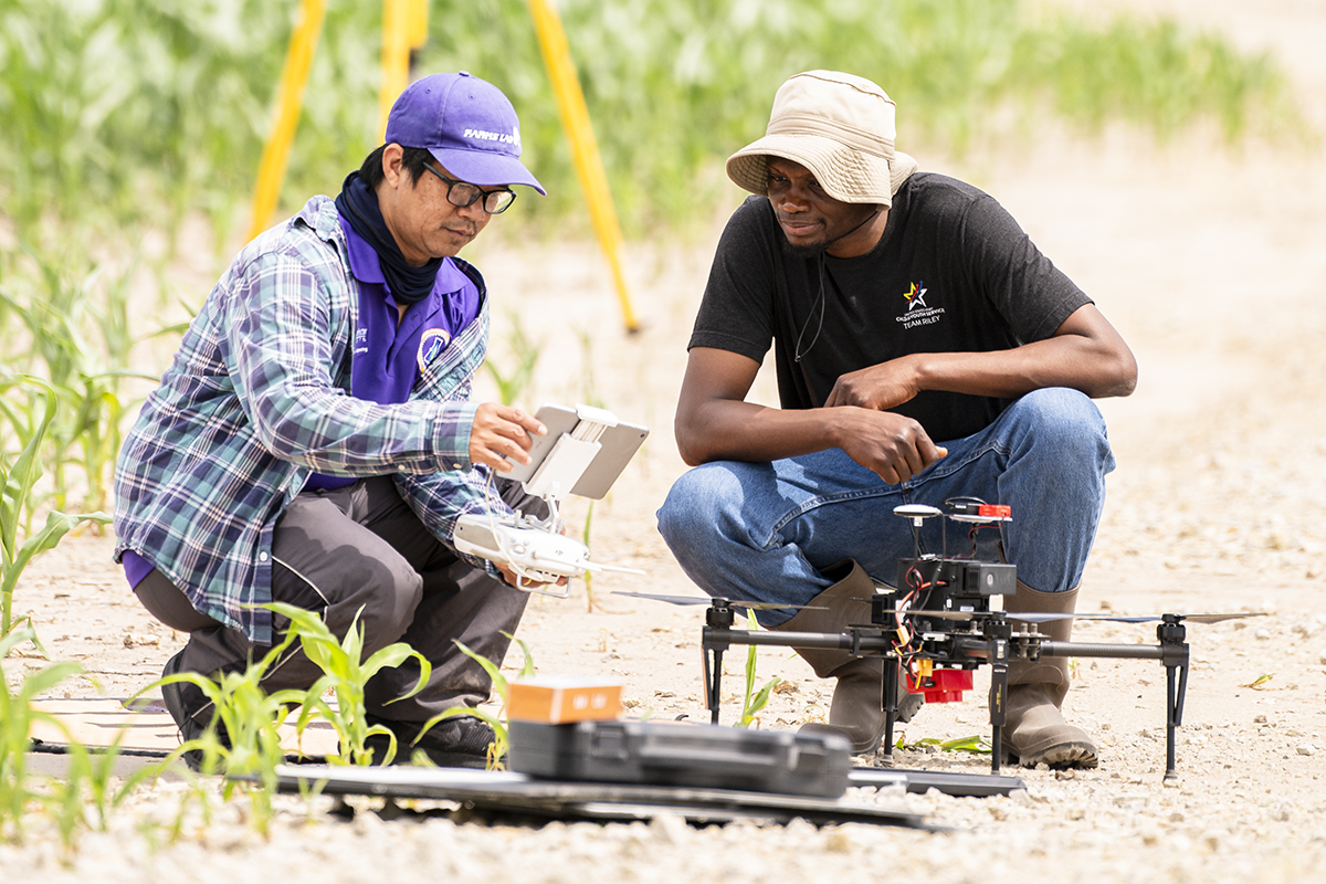 Two men crouch next to a drone sitting on dirt in a field of crops.