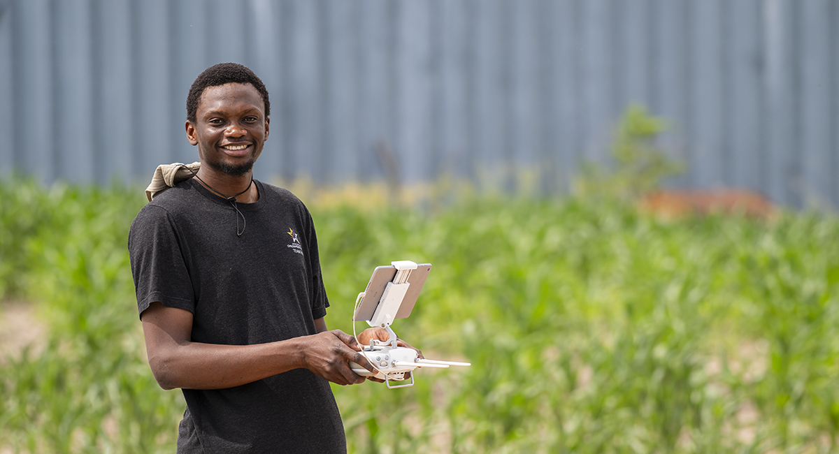 A doctoral student holds a drone controller and poses for a portrait in a field.