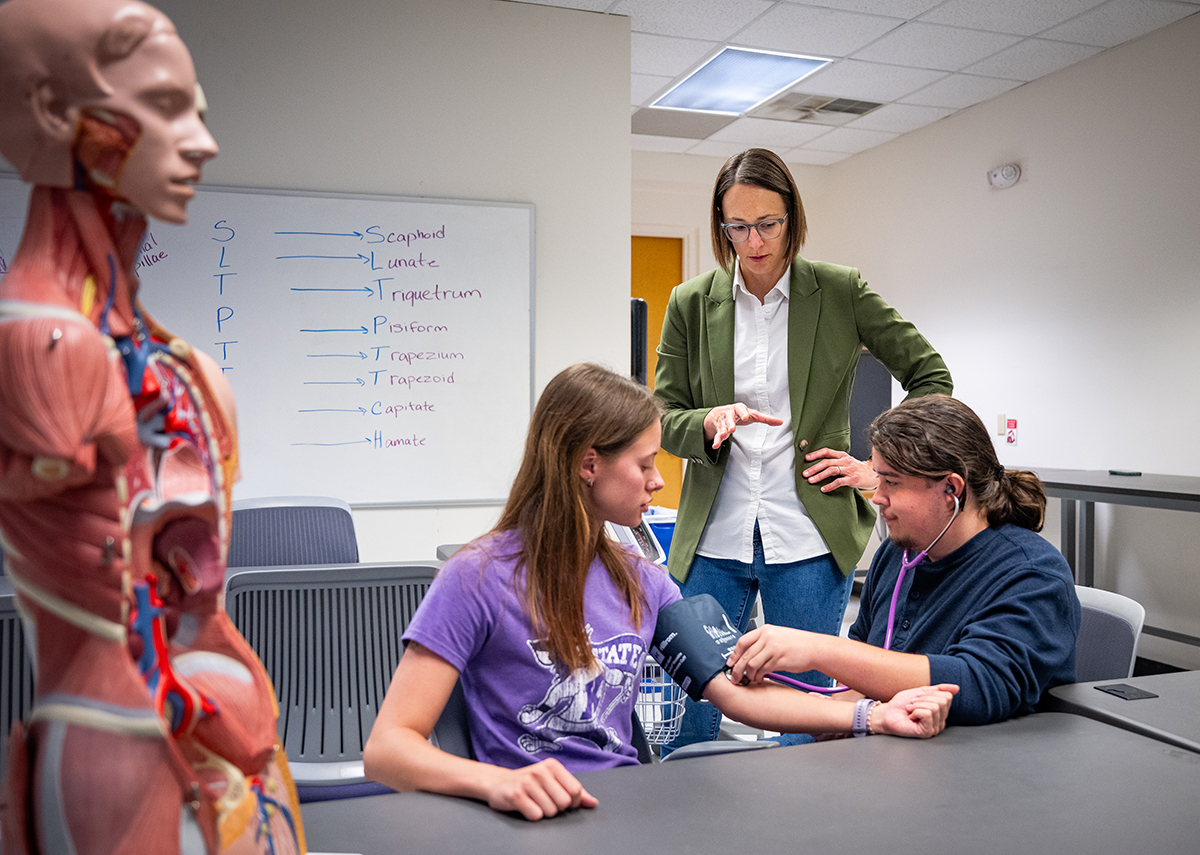 A woman in an olive green blazer instructs two students learning to take blood pressure.