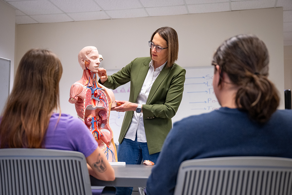 A woman stands next to a human body model and teaches students.