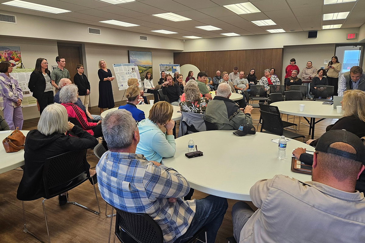 A group of adult community members sit in a large meeting area and hold a meeting.