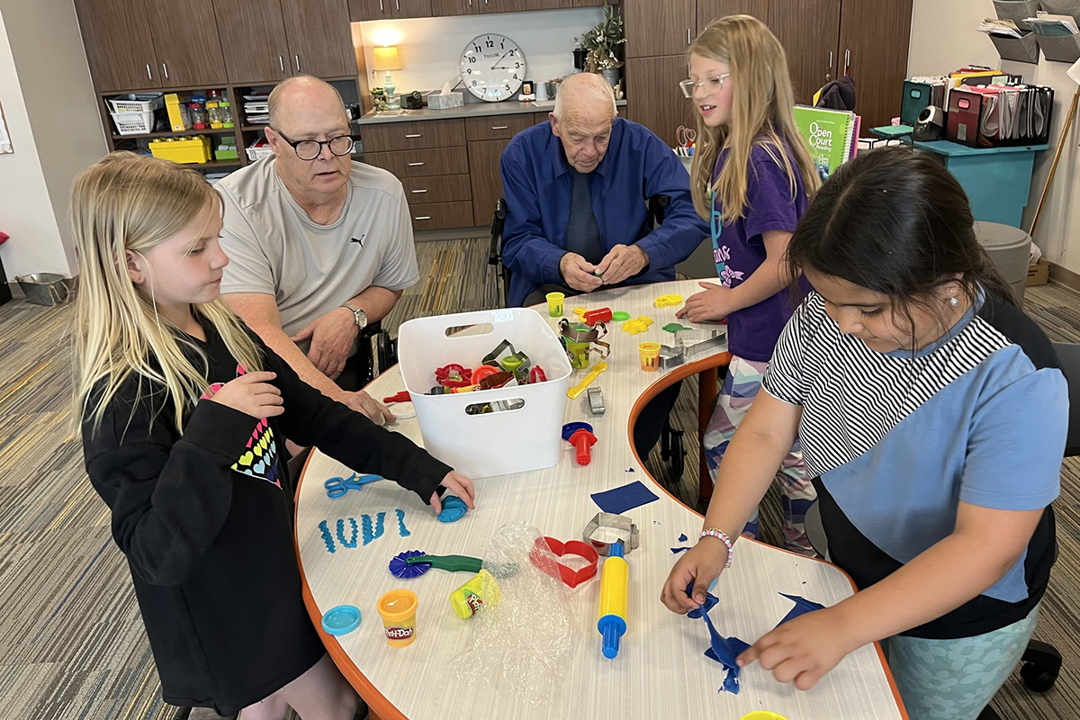 A group of elementary school children play with play dough alongside some senior citizens in a nursing home.