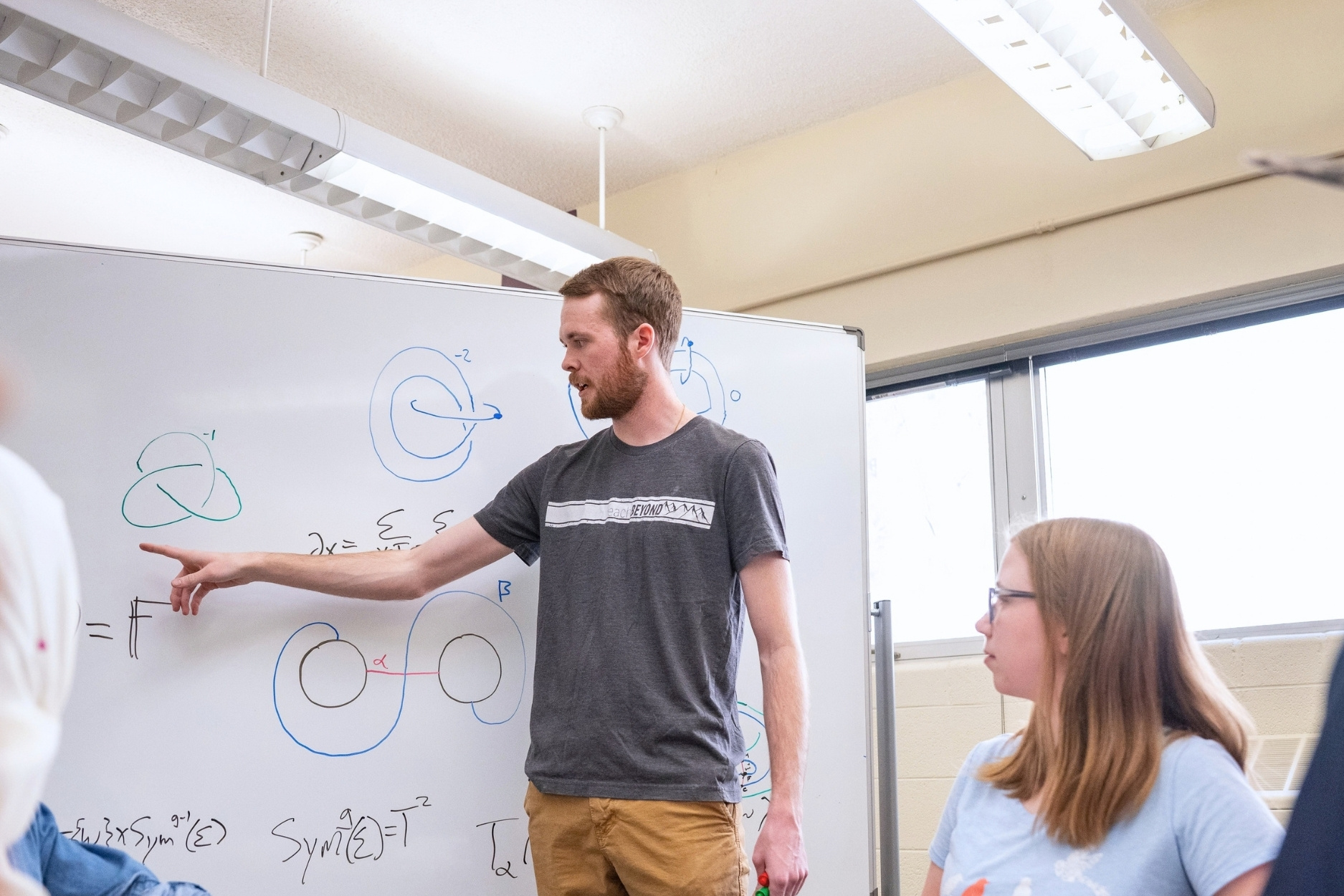 Jesse Osnes points at a whiteboard while explaining a mathematical solution to a table of college-aged students.
