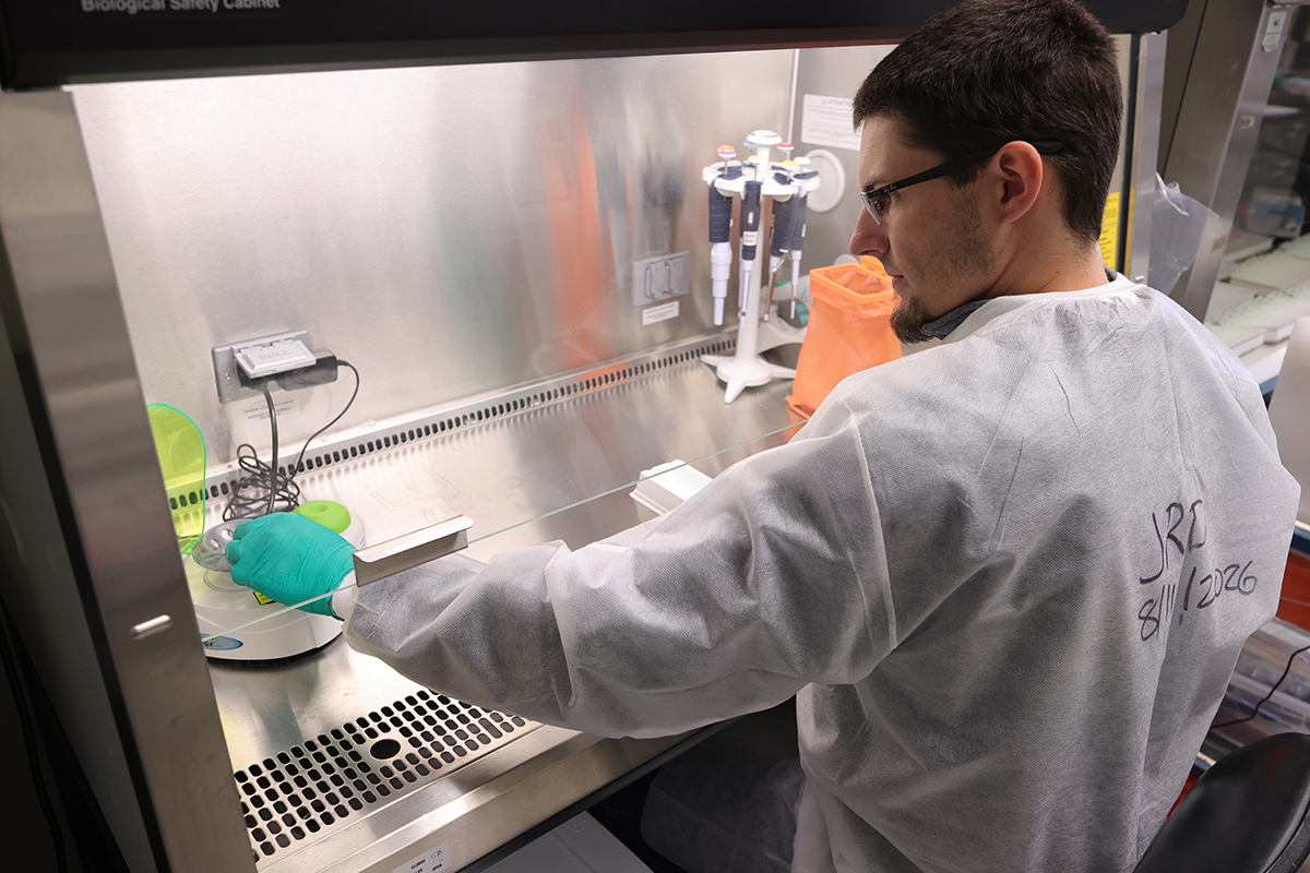 A student lab worker handles a sample and centrifuge in a laboratory equipment hood.