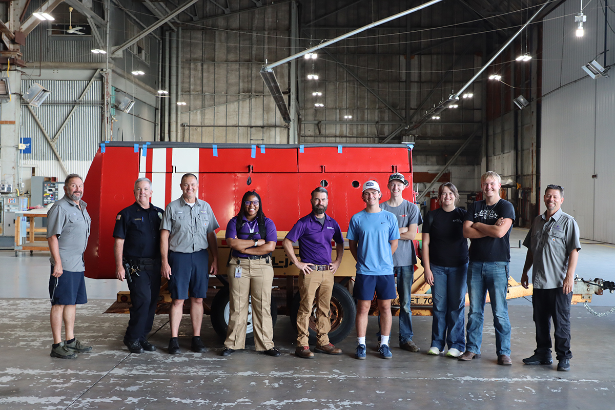 A group of 10 people stands in a row in front of a red metal plane wing on a trailer.